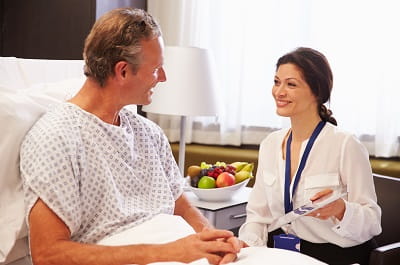 A female healthcare worker is seated while talking to a male patient who is in a hospital bed.