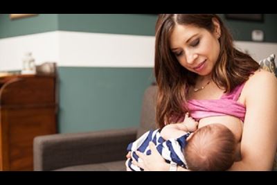 A young mother is breastfeeding her newborn while sitting on a couch at home.