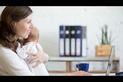 Young business woman holds her newborn while working on a laptop at home.