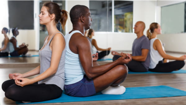 Group of positive people sitting back to back in lotus positions in modern yoga studio