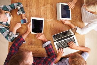A family is gathered around a table, and they all have digital devices.