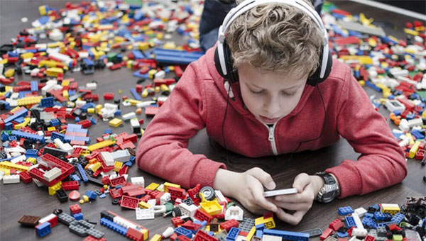 A boy is looking at his cellphone and listening to his headphones while surrounded by Lego bricks on the floor