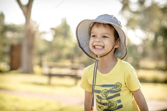 A smiling boy is wearing a large hat outdoors to protect his face from the sun