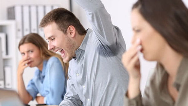 During a work meeting, a young man raises his left arm and reveals a sweat stain in the armpit area of his shirt. Two of his female coworkers are covering their noses.
