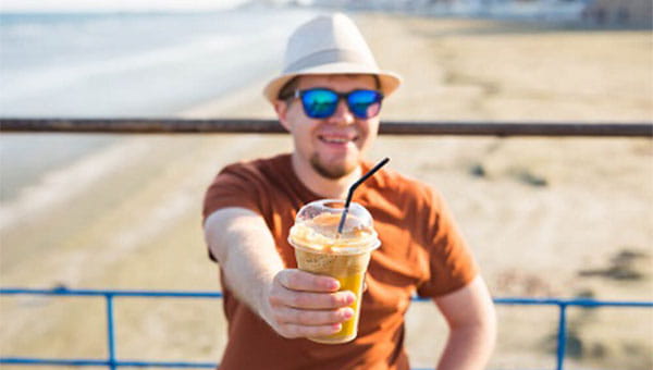 man wearing a hat and sunglasses on the beach holding a coffee drink