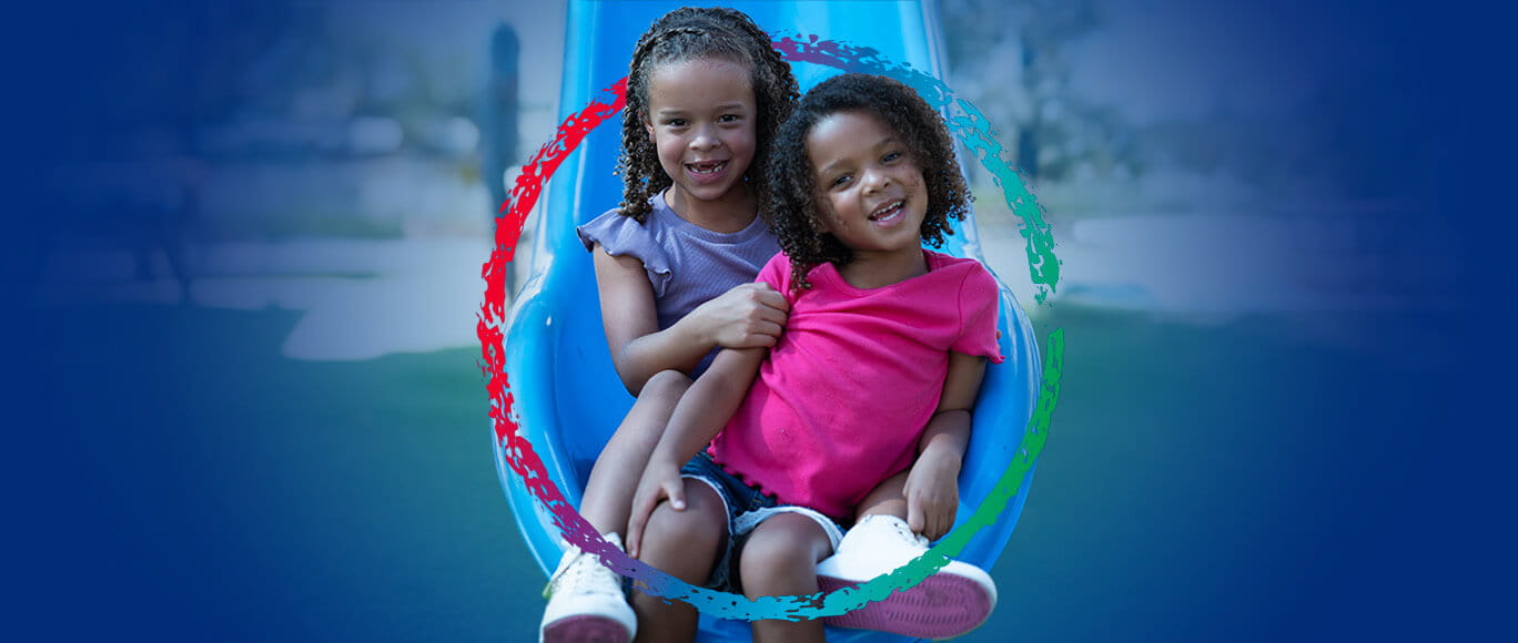 Two young black girls smiling together in a playground setting sitting at the end of a slide. They are surrounded by the BayCare Kids logo.