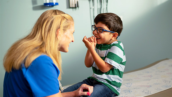A young boy with glasses sitting on an examination bed, laughing with a pediatric specialist in a medical setting.
