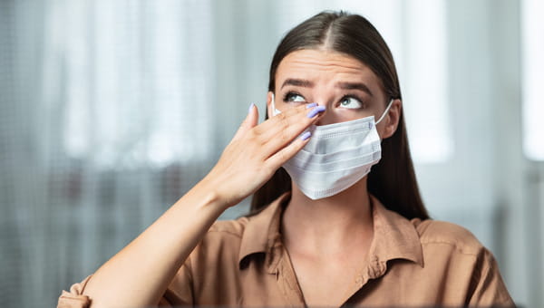 Girl wearing surgical mask rubbing her eye with dirty hands, working on laptop