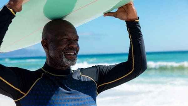 A black man stands next to the ocean while carrying a surfboard on his head.