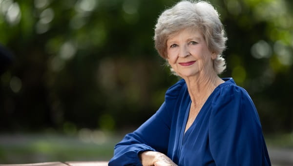 a woman with short gray hair wearing a long sleeve blue blouse sits outdoors with green foliage in the background
