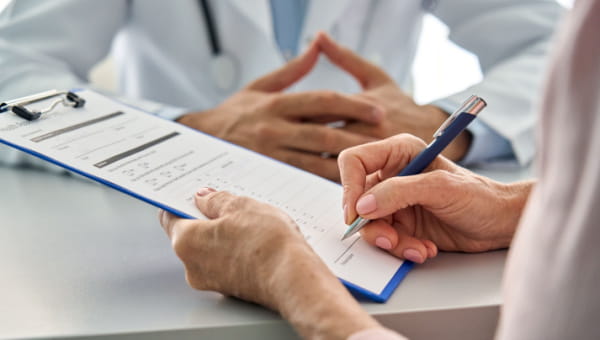The Why Behind Prior Authorizations Close up shot of older senior female patient hands holding pen filling medical heath data form sitting at desk with family doctor in modern hospital clinic