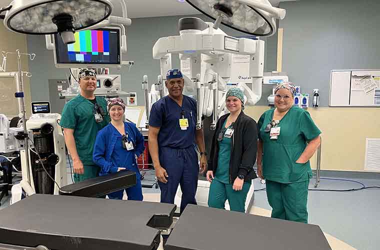 Five medical staff members in scrubs posing in a hospital operating room equipped with advanced medical machinery.
