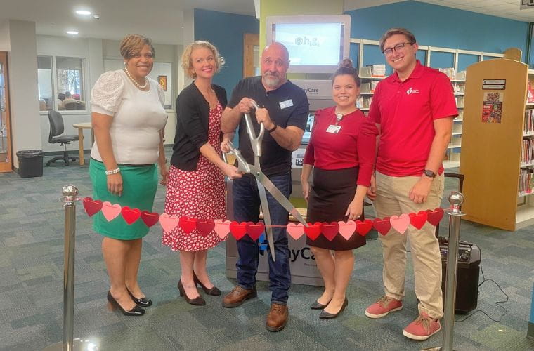 A group of five people are standing lined up in a library at a ribbon cutting event with a gentleman standing in the center holding an oversized pair of scissors.