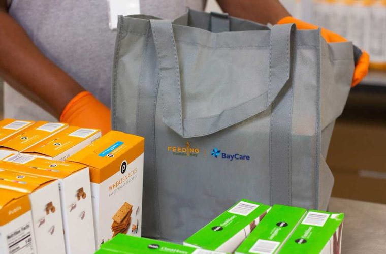A gray reusable bag with Feeding Tampa Bay and BayCare logos is shown surrounded by boxed crackers in green and white and orange and white boxes. An individual with a dark complexion stands behind the bag wearing neon orange gloves.