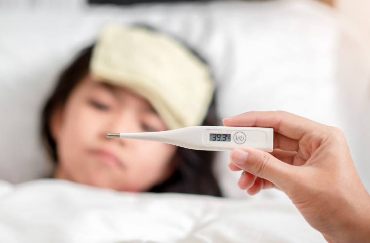 Mother holds digital thermometer with high temperature while daughter lays sick in bed with a cool towel on her forehead to reduce a high fever.