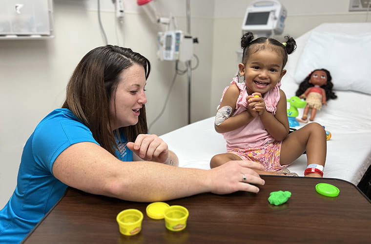 A female child life specialist entertains a young female patient who is smiling from her hospital bed.