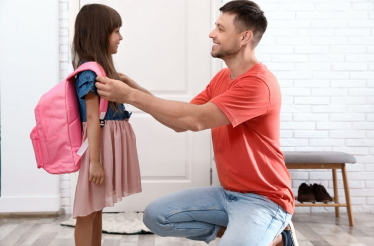 Happy father helps his little daughter put on her pink backpack at home.