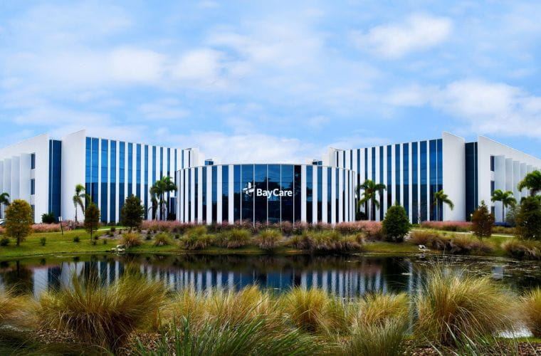 A modern office building is encircled by lush, tall grass, blending with a lake in front of it. The word BayCare is displayed on the front of the building. 