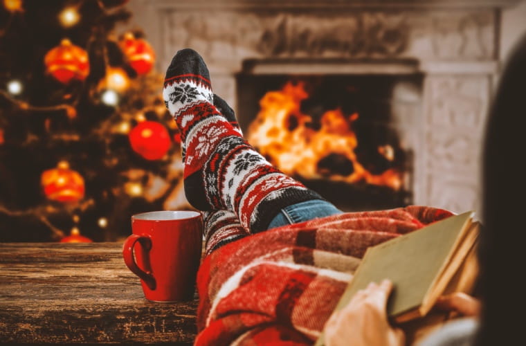 Person relaxing with cozy Christmas socks, legs propped on a coffee table, enjoying a warm drink and a book by a crackling fireplace.