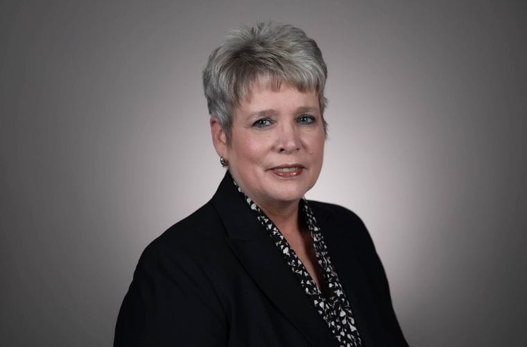 A headshot of Donna Lynch smiling. She is wearing a black jacket with a black and white floral print shirt underneath with small silver hoop earrings. She has very short, spiky salt and pepper color hair. 
