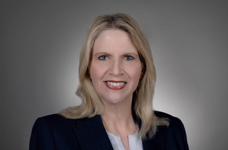 A headshot of Ann Marie Swindler smiling. She has below shoulder length straight blonde hair and is wearing a black jacket with a white shirt underneath. 