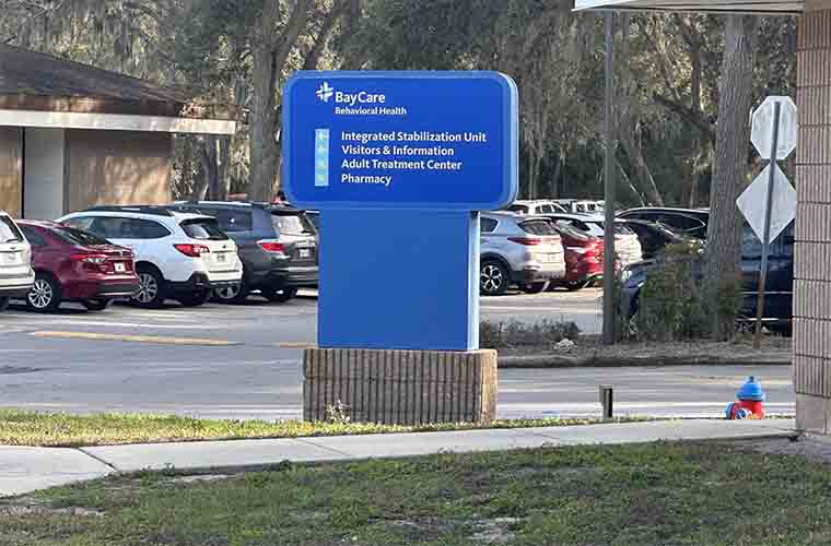 A blue directional sign in a parking lot with cars and other buildings.