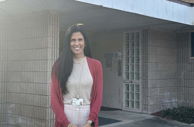 A woman with long dark hair stands in front of an older building on a sunny day.