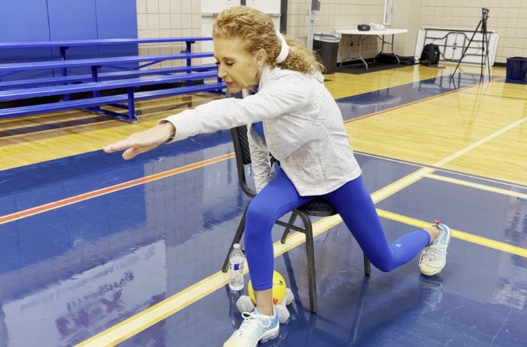 In a gym, a female fitness instructor with blue leggings on sits on a chair, with one arm stretched out.