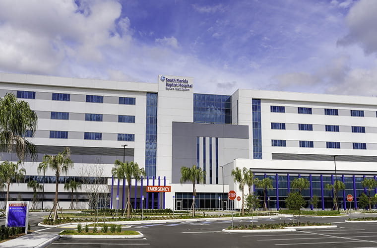 The exterior of South Florida Baptist Hospital. It’s a large hospital building against a blue sky with white clouds. Palm trees line the front of the building.