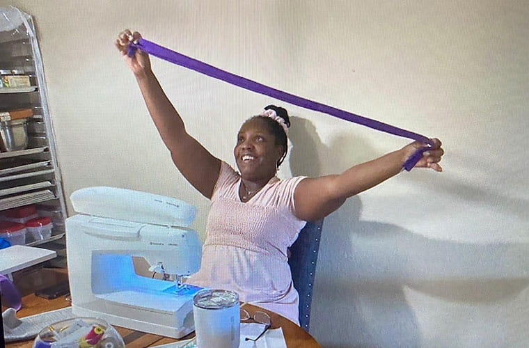 Black woman holds a long, narrow piece of purple fabric above her head. She gazes up with a huge smile. She is seated in front of a sewing machine. 