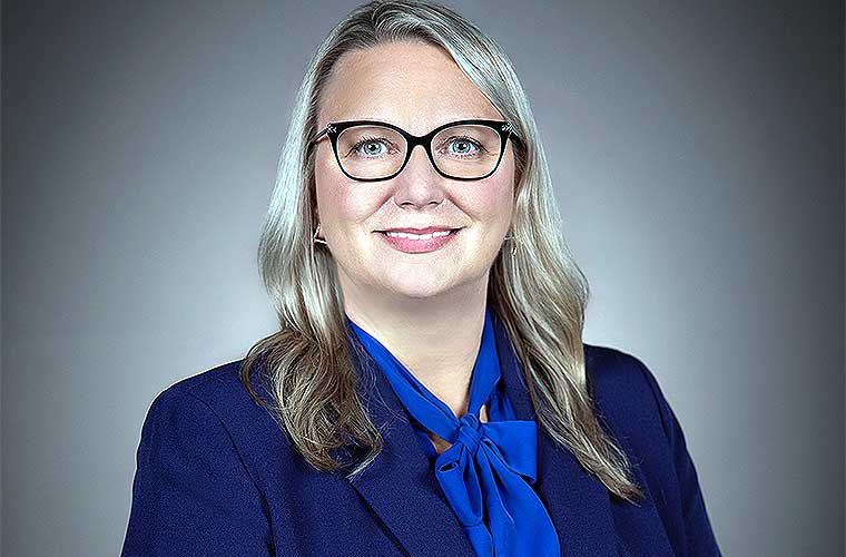A professional headshot of Lynnette Clinton smiling. She is wearing glasses, a navy jacket, a blue shirt with a bow tied in the front and she has wavy dark blonde hair.