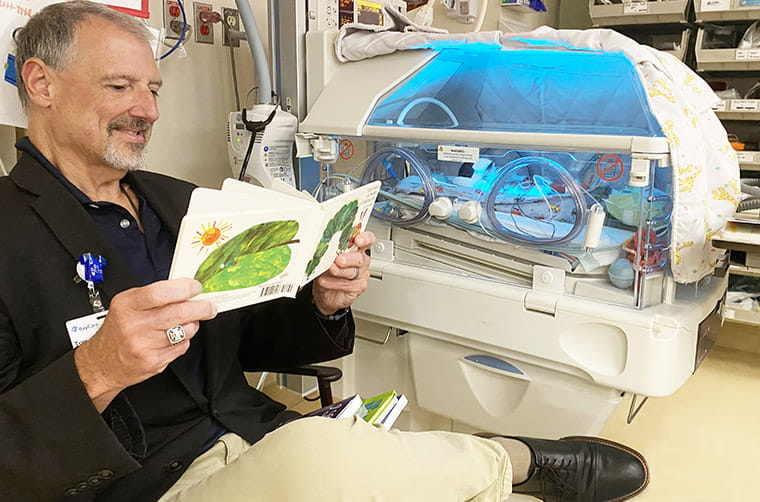 A man reads a book to a premature baby who is an isolette bed in a hospital room.