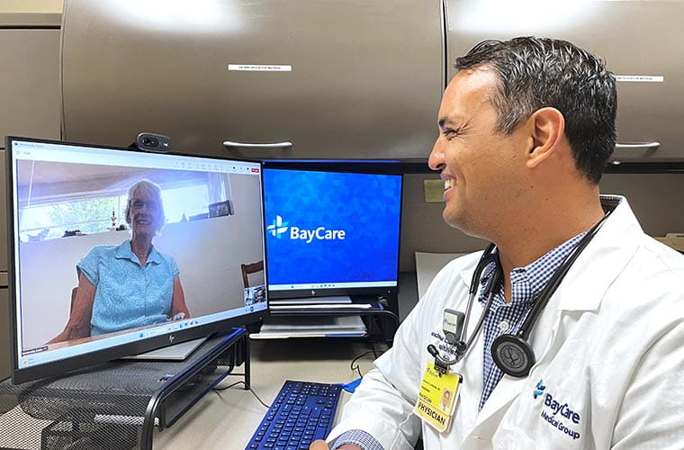 A doctor, wearing a stethoscope and a white doctor's coat with "BayCare Medical Group" embroidered on it, speaks with a person appearing via video conference on a computer screen. Both people are smiling, and a separate computer monitor in the background reads "BayCare."