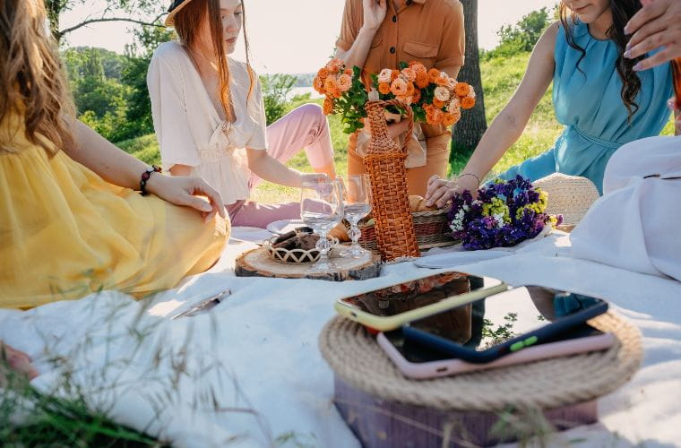 Friends gathered for a summer picnic, sharing food and laughter in a vibrant outdoor setting filled with sunshine and greenery. Their phones are placed aside.