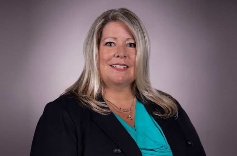A smiling headshot of Nanette Wilcox. She has short, below shoulder length straight blonde hair and is wearing a teal shirt with a black jacket and two layered gold necklaces.