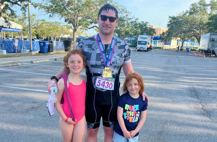 A man dressed in athletic clothing and wearing a race bib stands between a little girl dressed in a pink swimsuit and another wearing a T-shirt and light-blue shorts.
