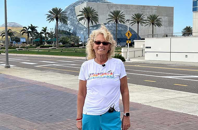 A woman wearing a T-shirt, shorts and sunglasses pauses in front of buildings.