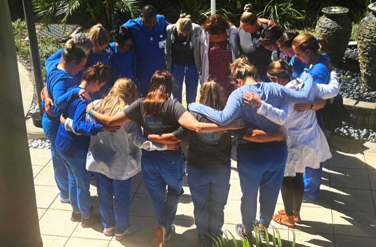 Seventeen BayCare nurse managers in blue scrubs stand in a circle in and outside courtyard with arms over each other's shoulders and heads bowed in prayer.