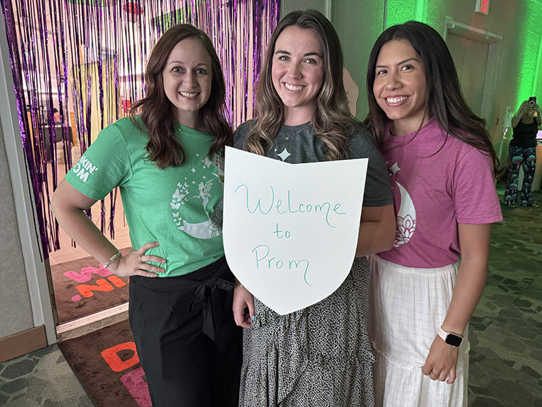 Three women in T-shirts and skirts proudly hold a welcome sign, smiling and standing together in a friendly pose.
