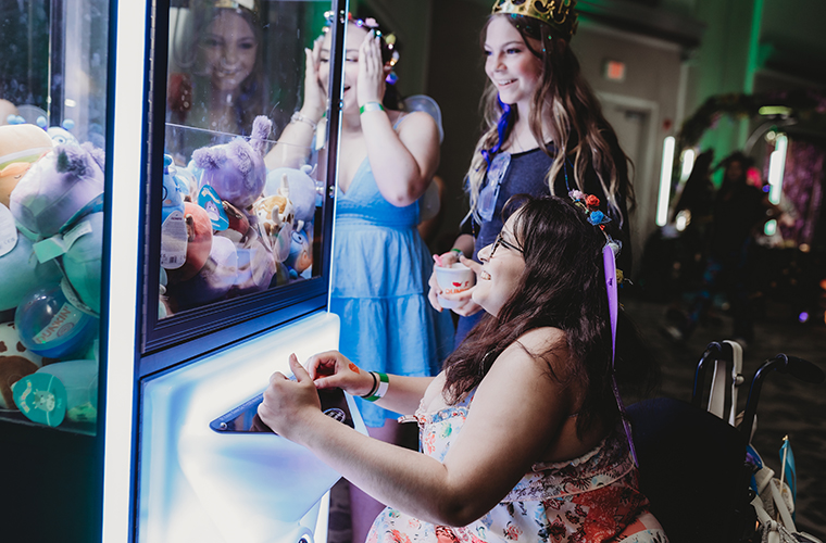 Three teenage girls wearing colorful dresses laughing in front of an arcade machine.