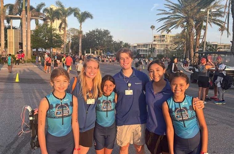 Three children wearing athletic clothing smile for the camera with three adults in a parking lot as they prepare for an athletic event.