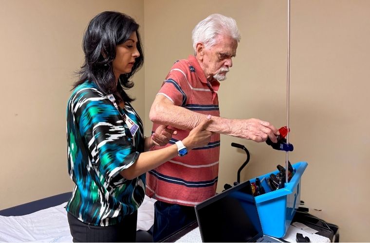 Woman with dark hair assists an elderly man with the use of his arm in a clinical office.