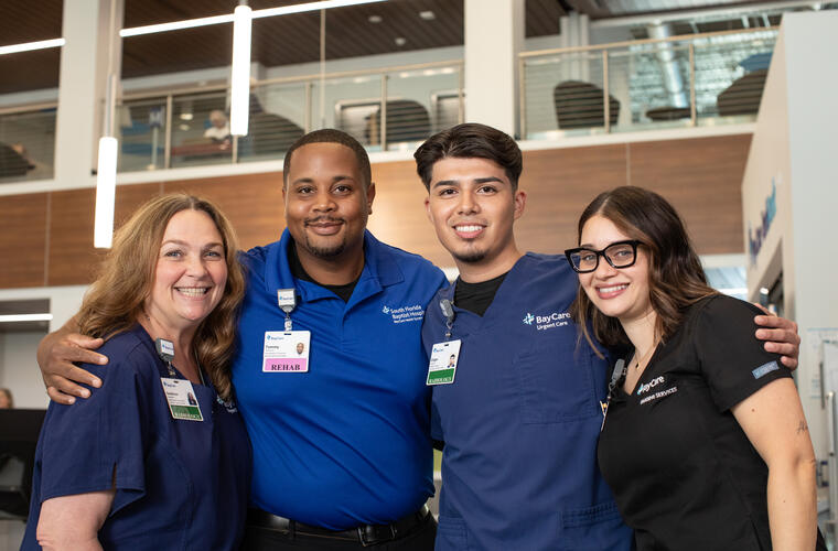 A diverse group of BayCare team members wearing work scrubs stand smiling in front of a BayCare facility.
