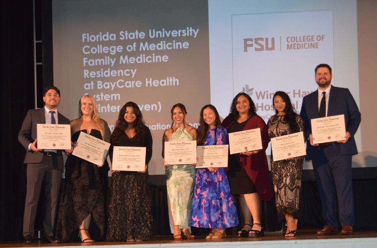 Eight people stand on a stage holding their diplomas during a graduation ceremony.