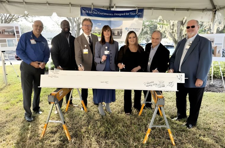 A group of seven people stand behind a construction beam and smile for a photo.