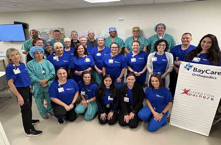 Group of medical professionals in scrubs, standing and kneeling, posing with a "BayCare Orthopedics Operation Walk USA" banner in a clinical setting.