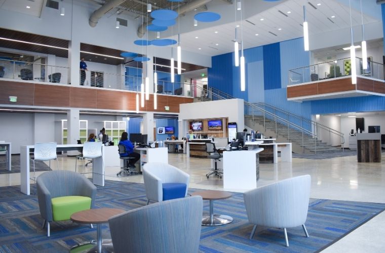 Spacious, modern lobby of BayCare HealthHub (Bloomingdale) featuring high ceilings with exposed ducts, hanging circular blue acoustic panels, and vertical blue accent walls. The area includes a central reception desk with a sign reading ‘BayCare HealthHub,’ polished concrete floors, and patterned blue carpet sections. There are seating areas with chairs on the right, a staircase leading to an upper level with glass-front offices and abundant natural light streaming through large windows.