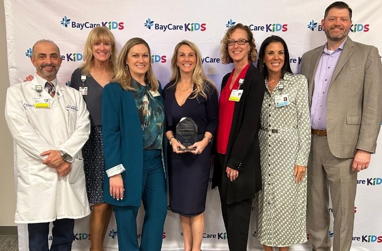 A group of male and female professionals pose in front of a BayCare Kids banner while the woman in the middle is holding a plaque, honoring her as FHA Caregiver of the Year.