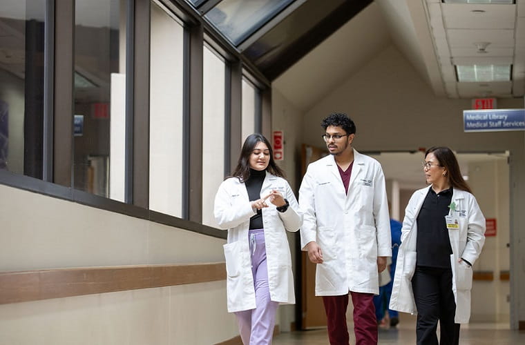 Two graduate medical students – one female, one male – with a female physician walking down a hospital hallway; female has long dark hair and is wearing an open white lab coat, black turtleneck and lavender scrub pants; male is taller with short curly hair, glasses and is wearing a closed white lab coat over burgundy scrubs; female physician has long dark hair, glasses and is wearing an open white lab coat with black scrubs and an attending physician badge.