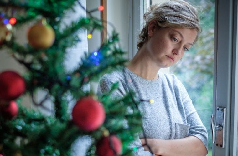 A woman looking down sadly poses next to a festive Christmas tree adorned with lights and ornaments.